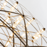 Pendant light close-up shows intersecting metal strips and glowing clear tubes with gold-toned ends against a pale background.