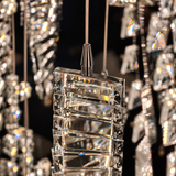 A close-up of a modern crystal chandelier pendant with a faceted clear glass block and silver metal rod, surrounded by other hanging crystal elements against a dark background.