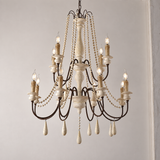 A multi-tiered chandelier with a distressed white wood finish and dark metal arms, decorated with hanging pearl bead strands and teardrop crystal drops, shown lit against a plain light gray wall.