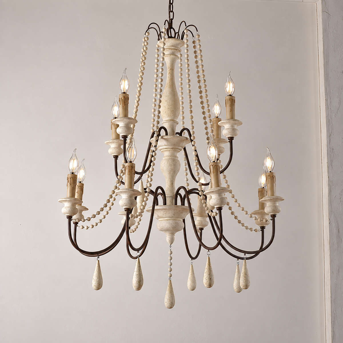 A multi-tiered chandelier with a distressed white wood finish and dark metal arms, decorated with hanging pearl bead strands and teardrop crystal drops, shown lit against a plain light gray wall.