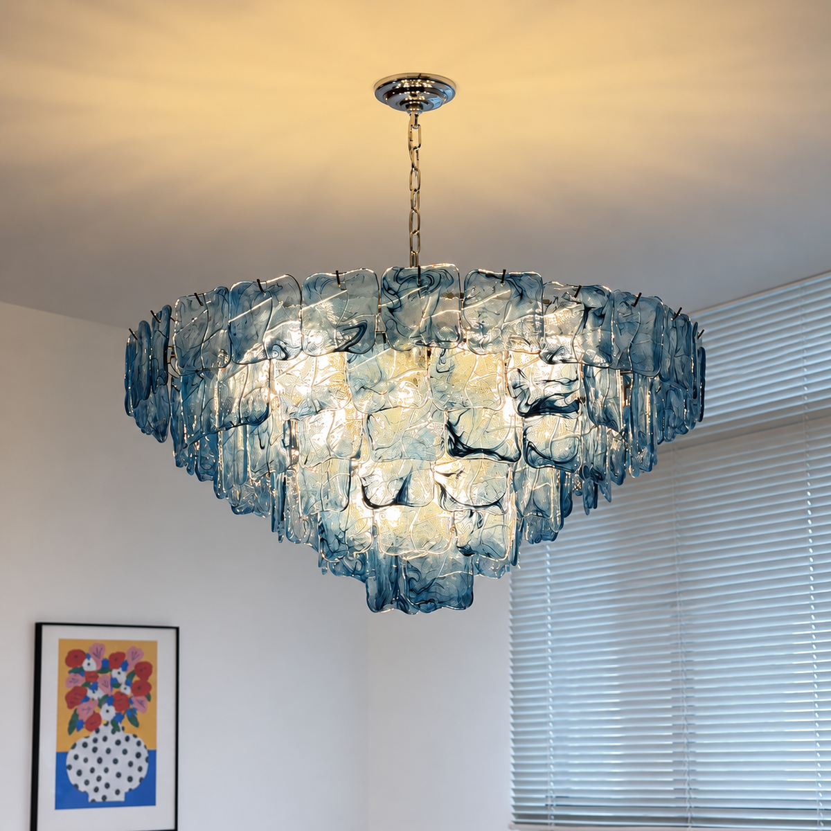 A large, tiered chandelier made of textured, translucent blue glass pieces, suspended from a gold-toned chain and ceiling canopy in a room with white walls and window blinds.