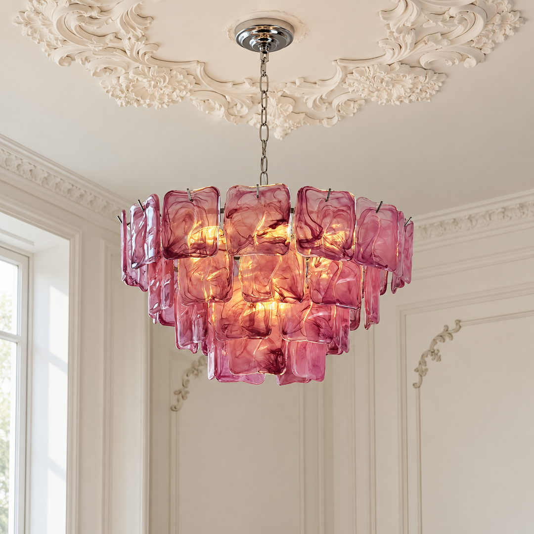 A pink glass chandelier with a tiered, blocky design hangs from a silver chain in an elegant room with white decorative ceiling moldings and a window.