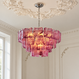 A pink glass chandelier with a tiered, blocky design hangs from a silver chain in an elegant room with white decorative ceiling moldings and a window.