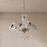 A clear glass chandelier with multiple bell-shaped shades and hanging teardrop crystals, suspended from a chain against a textured beige wall and ceiling.