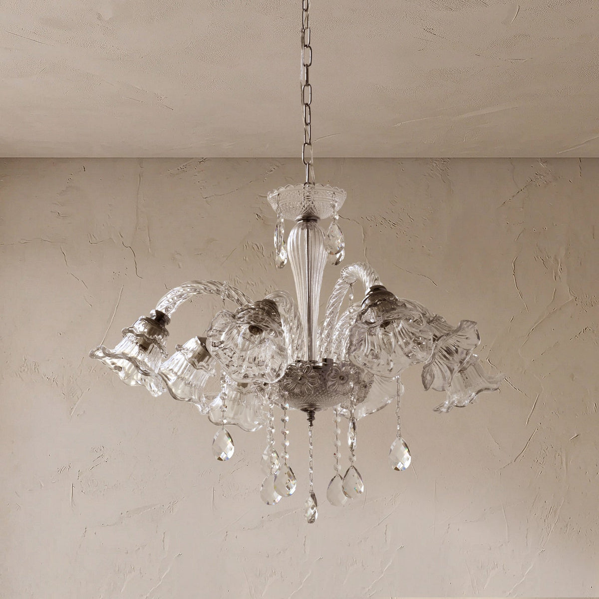 A clear glass chandelier with multiple bell-shaped shades and hanging teardrop crystals, suspended from a chain against a textured beige wall and ceiling.