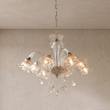 A clear glass chandelier with flower-shaped shades, glowing warm bulbs, and crystal teardrop pendants hangs from a chain against a textured beige wall.