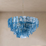 A modern chandelier featuring multiple tiers of textured, translucent blue glass blocks suspended from a central chain, set against a neutral beige plaster wall and ceiling.