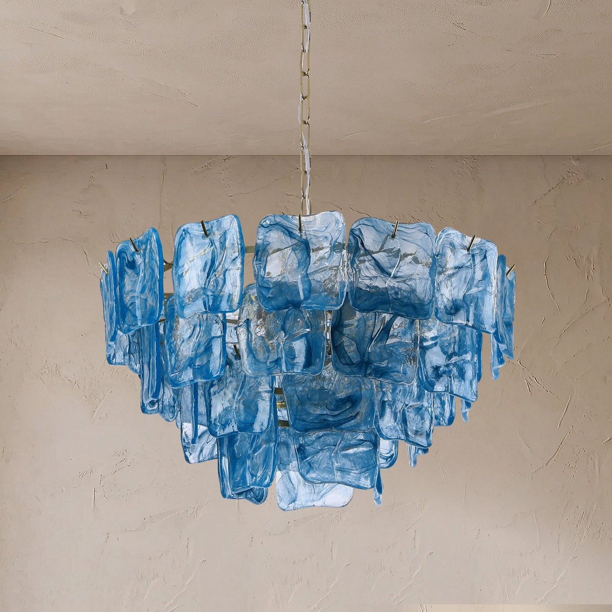 A modern chandelier featuring multiple tiers of textured, translucent blue glass blocks suspended from a central chain, set against a neutral beige plaster wall and ceiling.