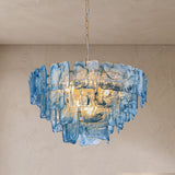 A modern chandelier pendant light made of layered translucent blue glass pieces, suspended by a gold-toned chain against a textured beige plaster wall and ceiling.