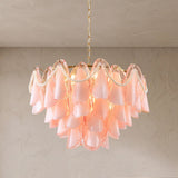 A modern tiered chandelier with layered blush pink bell-shaped glass shades, accented by white pearl bead garlands and warm white lights, hanging from a gold chain against a textured beige wall.