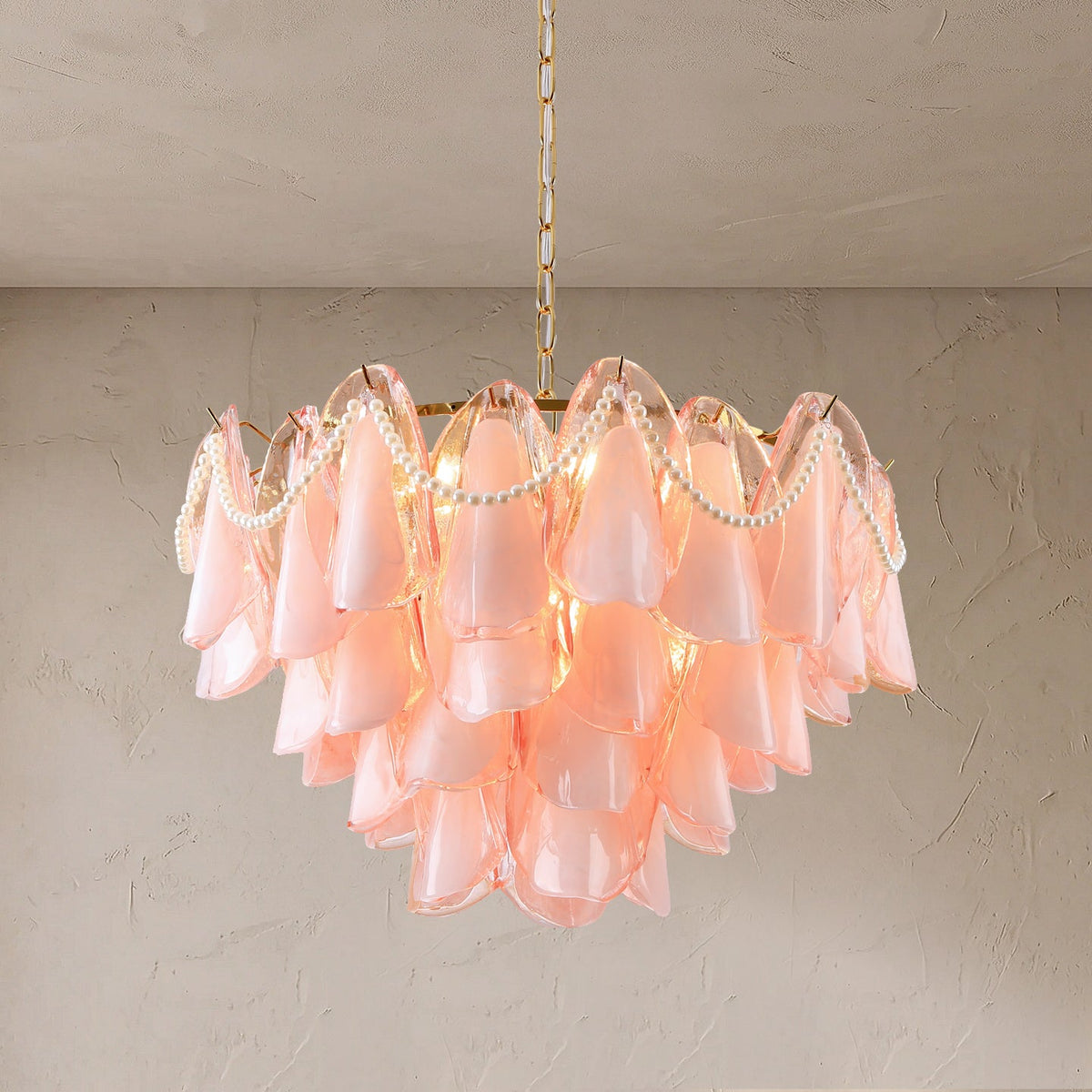A modern tiered chandelier with layered blush pink bell-shaped glass shades, accented by white pearl bead garlands and warm white lights, hanging from a gold chain against a textured beige wall.