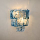 Illuminated wall sconce with three tiers of rippled blue glass panels casting warm light onto a beige wall.