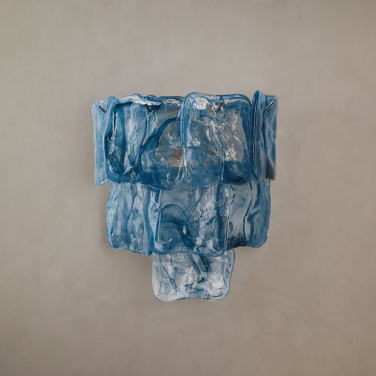 Blue glass wall sconce with three tapering tiers of rippled panels against a beige wall.
