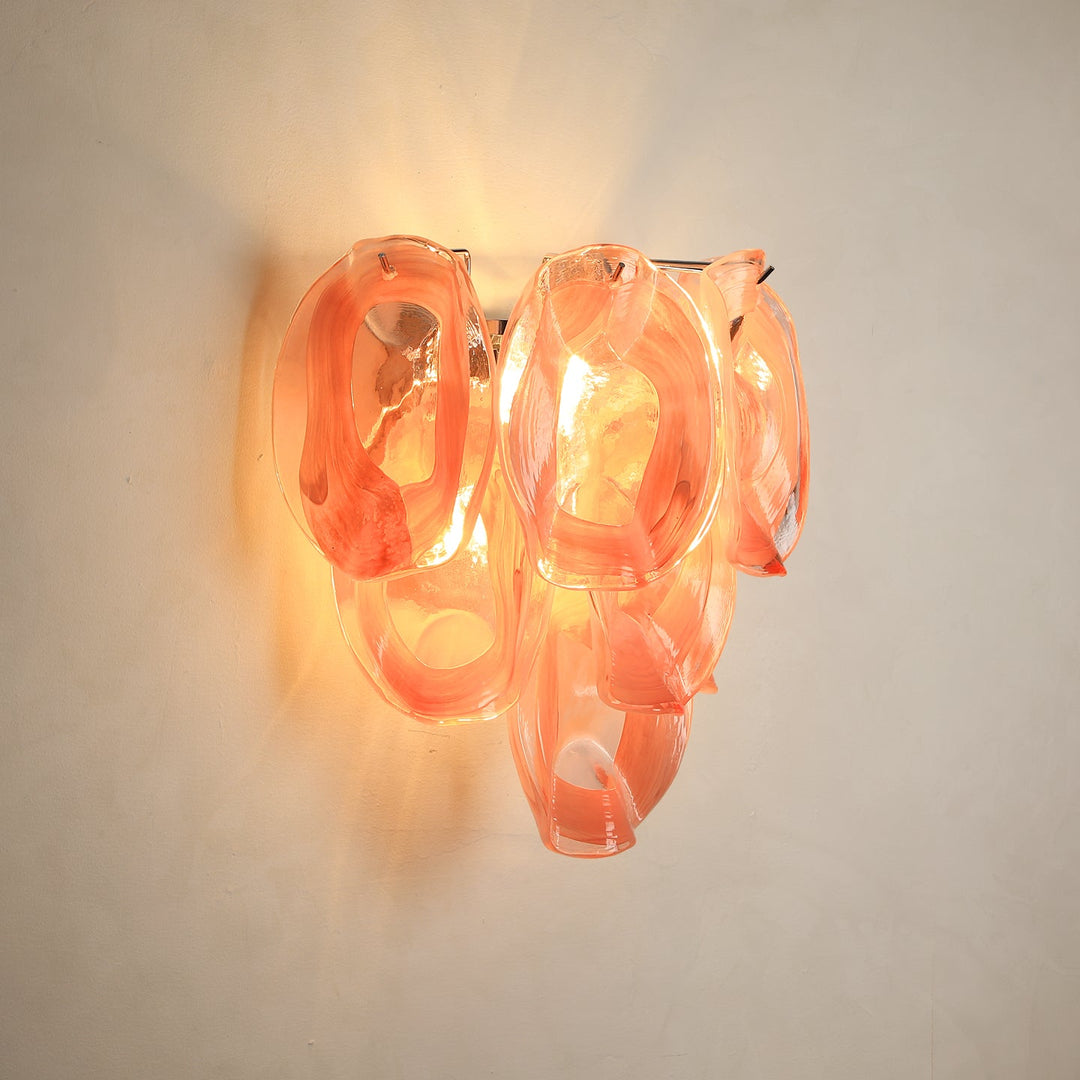 A modern wall sconce light fixture with organic, flowing translucent glass shapes in a warm peach-orange color, casting a soft glow against a plain off-white wall.