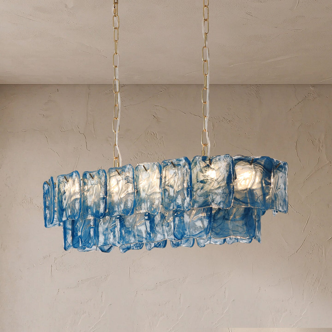 An illuminated linear chandelier with layered blue textured glass panels hangs from gold-toned chains against a beige textured wall.