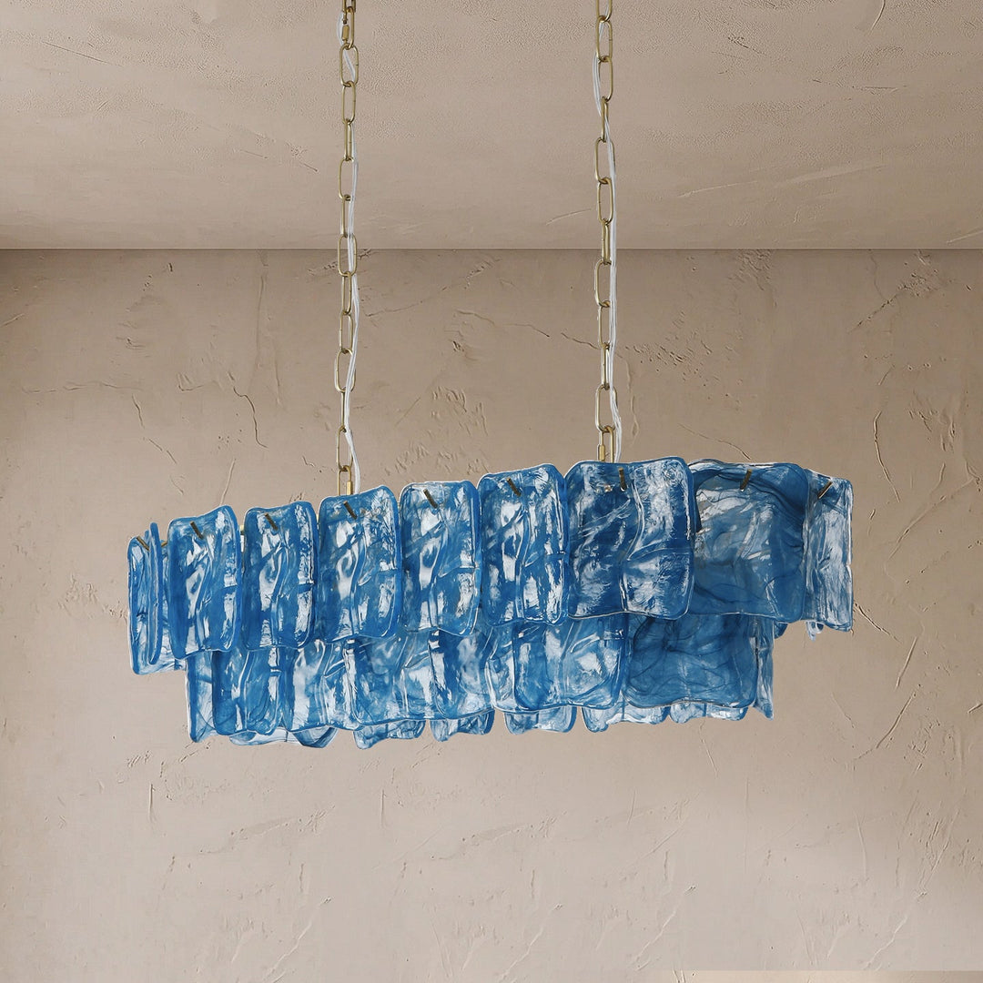 An unlit linear chandelier with layered blue textured glass panels hangs from two chains against a beige textured wall.