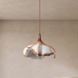 A modern pendant light with a sculptural, flower-like shade made of layered white, beige, and copper-toned petals, suspended by a thin copper cord against a textured beige plaster wall.