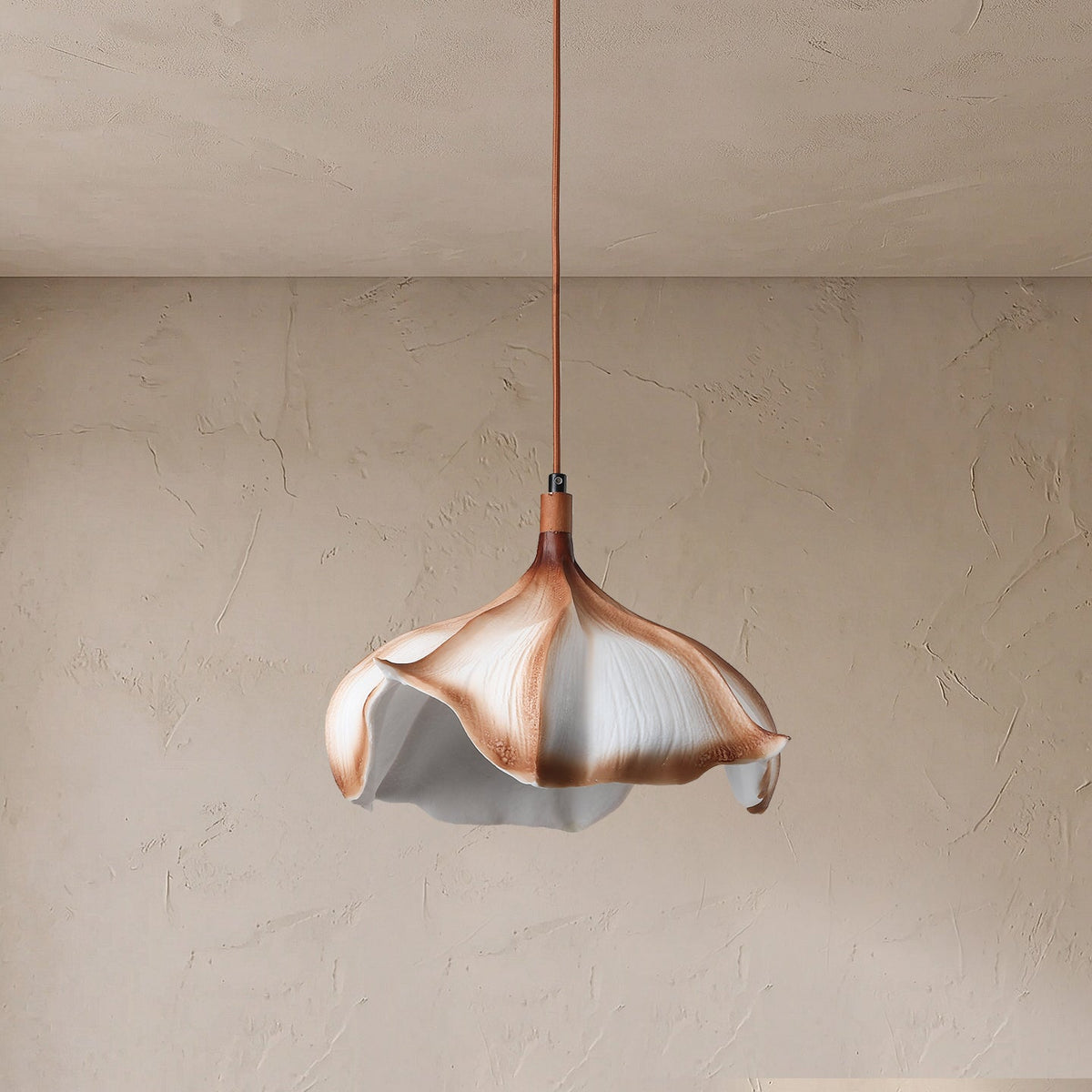A modern pendant light with a sculptural, flower-like shade made of layered white, beige, and copper-toned petals, suspended by a thin copper cord against a textured beige plaster wall.