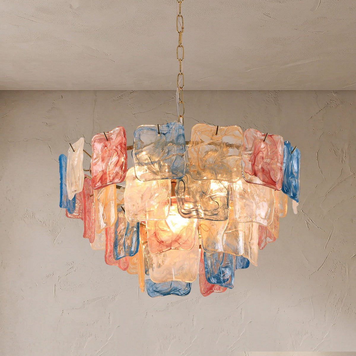 A modern chandelier with multiple layers of translucent, hand-blown glass panels in blue, pink, and peach tones, suspended from a chain against a textured beige wall and ceiling.