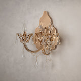 A two-light wall sconce with a polished gold-tone metal frame and clear crystal hanging drops, mounted on a textured light gray plaster wall.