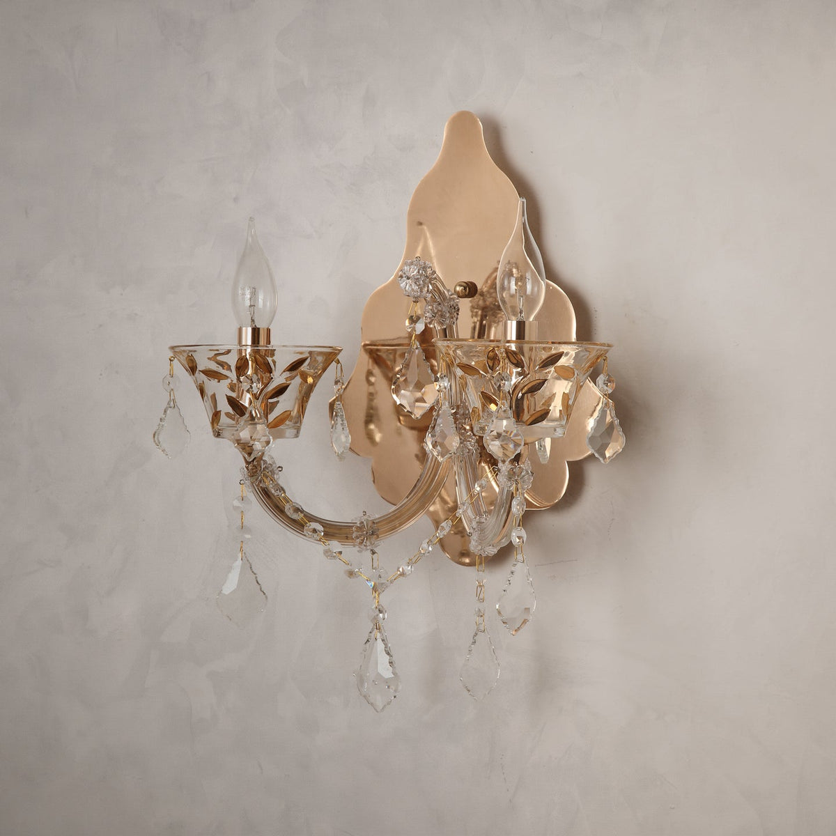 A two-light wall sconce with a polished gold-tone metal frame and clear crystal hanging drops, mounted on a textured light gray plaster wall.