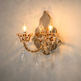 A two-light wall sconce with ornate gold-toned metalwork and clear crystal prisms, shown lit against a textured beige wall.