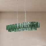 An unlit linear chandelier with two tiers of rippled green glass panels hangs from silver-toned chains against a beige wall.