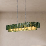 An illuminated linear chandelier with rippled dark green glass panels hangs from silver-toned chains against a textured beige wall.