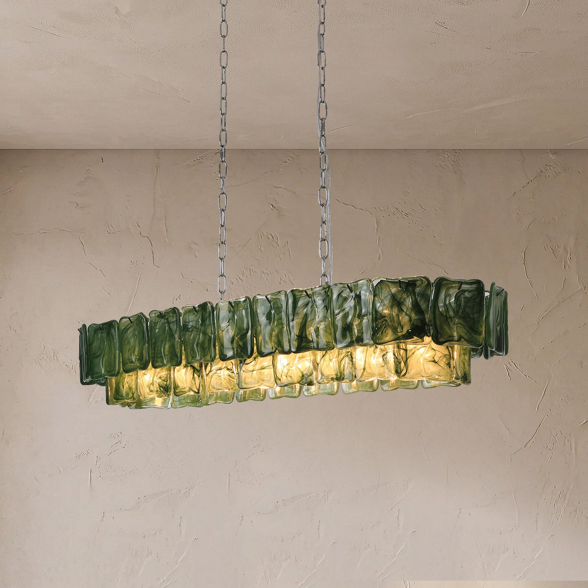 An illuminated linear chandelier with rippled dark green glass panels hangs from silver-toned chains against a textured beige wall.
