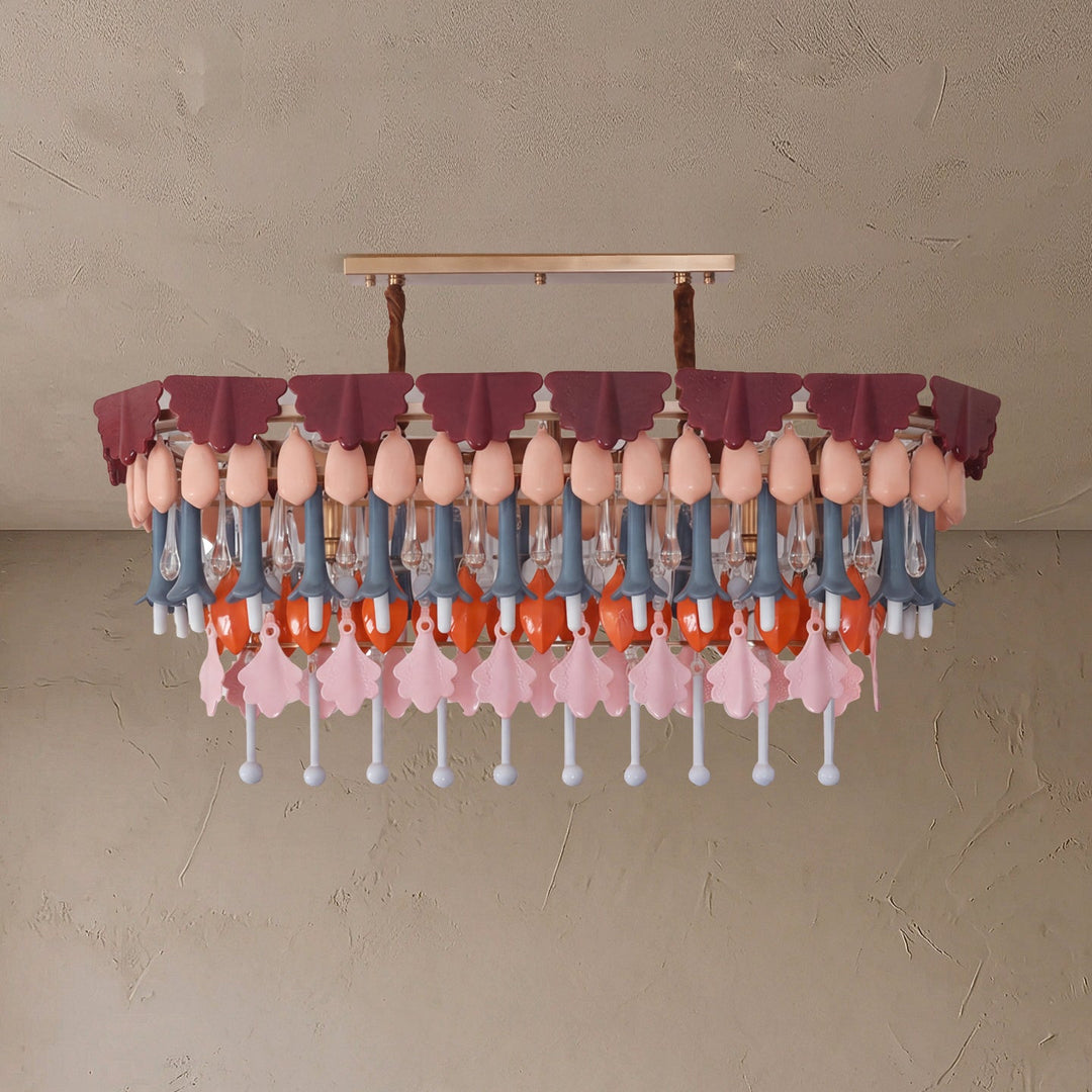 A modern chandelier with a tiered design, featuring dusty rose fabric ruffles, peach-colored bulb-shaped accents, blue-gray and orange hanging elements, and pink flower-shaped pendants against a textured beige wall.