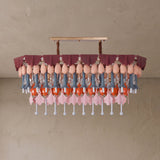 A modern chandelier with a tiered design, featuring dusty rose fabric ruffles, peach-colored bulb-shaped accents, blue-gray and orange hanging elements, and pink flower-shaped pendants against a textured beige wall.