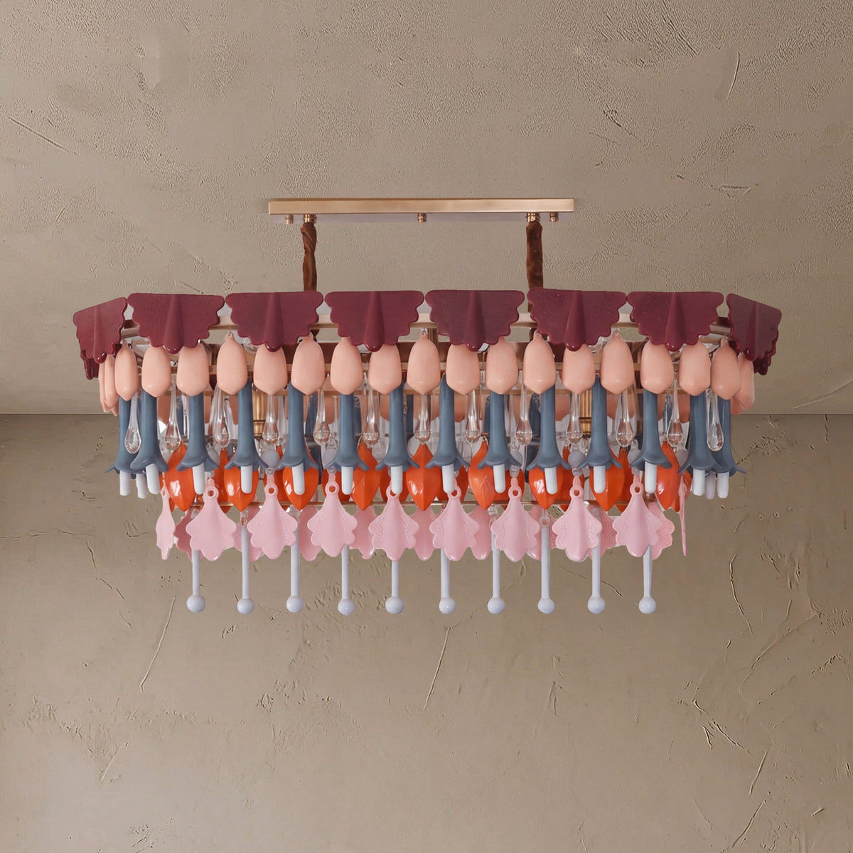 A modern chandelier with a tiered design, featuring dusty rose fabric ruffles, peach-colored bulb-shaped accents, blue-gray and orange hanging elements, and pink flower-shaped pendants against a textured beige wall.