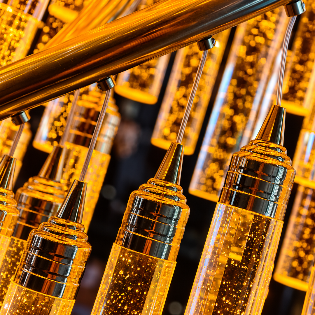A close-up of a modern chandelier featuring multiple clear cylindrical glass or acrylic pendant tubes with warm yellow illuminated internal lights and polished metallic silver and gold-toned caps, suspended from a thick gold-toned horizontal bar.