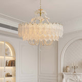 An elegant chandelier with a gold-tone metal frame and cascading white lace-like petals, adorned with hanging pearl beads, suspended from a chain in a bright, classic white room.