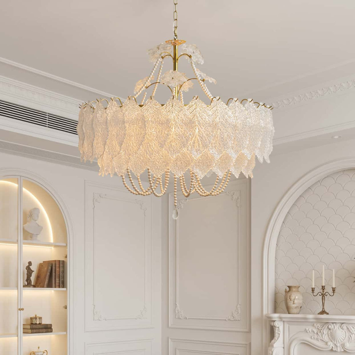An elegant chandelier with a gold-tone metal frame and cascading white lace-like petals, adorned with hanging pearl beads, suspended from a chain in a bright, classic white room.