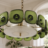 A modern chandelier with textured olive green glass sunburst discs and a clear glass bowl shade, suspended by a brass chain in a bright, elegant room.