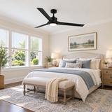 A modern black ceiling fan with a light fixture is mounted on a white ceiling in a beautifully styled bedroom with beige walls, a bed with blue and cream bedding, and a patterned blue rug.