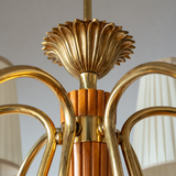 Close-up detail of a brass chandelier with curved arms, a fluted central column, and a decorative top finial, with soft-focus white lampshades in the background.