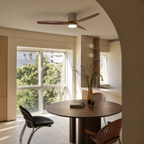 Ceiling fan light with three dark wood blades and a silver housing above a round dining table in a sunlit room.