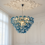 A blue and clear glass chandelier with a sculptural, wavy bubble design hangs from a silver chain in an elegant room with white paneled walls and a decorative ceiling medallion.