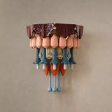 A multicolored ceramic and glass wall sconce with a burgundy top, peach shades, and teal, orange, and blue hanging drops on a beige wall.