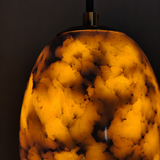 A close-up of a hanging pendant light shade made from a polished agate or marble-like stone, glowing warm amber-orange with dark brown and black natural veining against a dark background.
