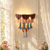 A decorative wall sconce with a warm glowing light, featuring dark brown leaf-like top pieces, peach and blue flower-shaped accents, and orange hanging details, mounted on a cream paneled wall between sheer white lace curtains.