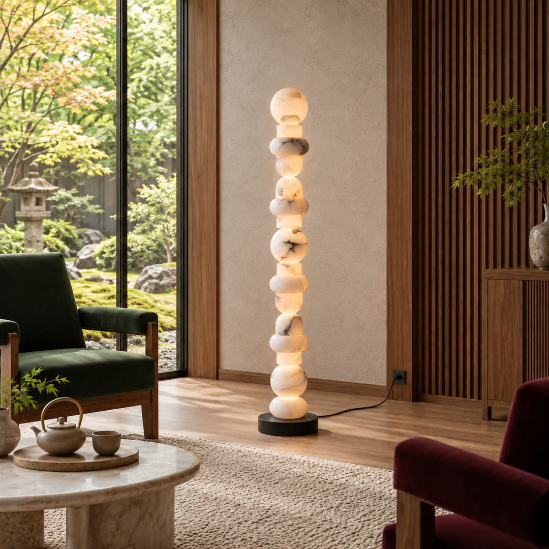 A tall, modern floor lamp made of stacked glowing stone spheres on a black base, placed in a styled living room with wood slats, chairs, and a window view of a garden.