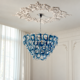 A blue and clear glass chandelier with a tiered, layered design hangs from a decorative white ceiling medallion in a bright, elegant room with white paneled walls and sheer curtains.