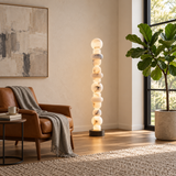 A modern floor lamp with a stacked, glowing marble sphere design stands in a sunlit living room next to a brown leather armchair and a large potted fiddle-leaf fig plant.