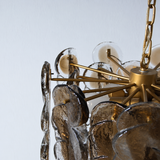 Close-up of a hanging chandelier with a brass-toned metal frame, clear glass globes, and layered translucent amber-gray glass discs, shown against a plain light wall.