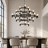 A modern multi-tier black metal chandelier with illuminated candle-style bulbs hangs above a dark wood dining table and upholstered chairs in a bright dining room with a white brick wall.