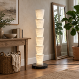 A modern floor lamp with seven stacked, tapered white textured shades on a round black base, styled in a warm, rustic living room next to a wooden console table, a tall mirror, and potted plants.