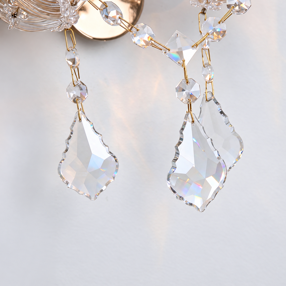 Close-up of a glass wall sconce with clear faceted crystal drops and gold-tone links against a pale gray wall.